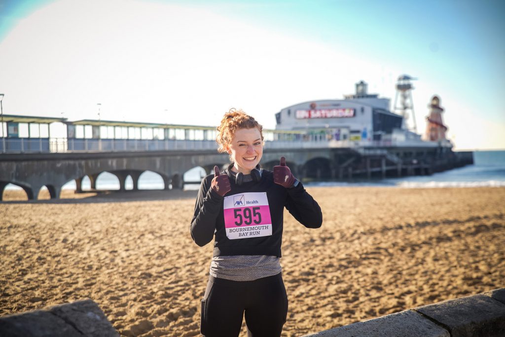 Woman runner with her thumbs up smiling at the camera next to Bournemouth beach 